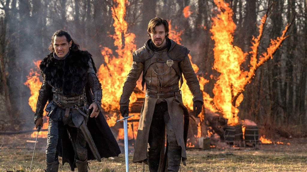 Two characters from The Outpost standing in front of a burning building
