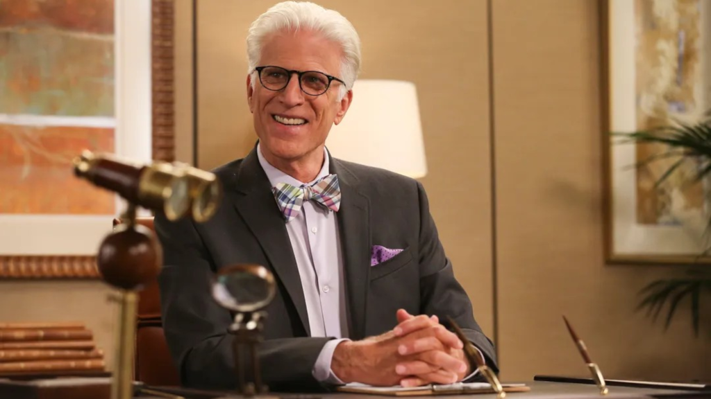 Michael sitting at a desk and smiling in The Good Place