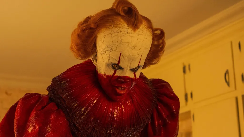 Bill Skarsgard's Pennywise smiles covered in blood standing in a kitchen from It Welcome to Derry